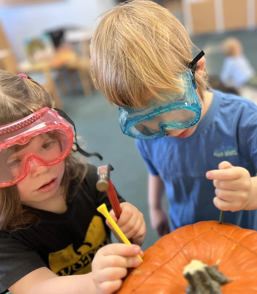 Two children, one wearing red work goggles and the other wearing blue, stand over a pumpkin. They are both focused on fine motor skills, carefully holding golf pegs and using small hammers to gently nail the pegs into the pumpkin.